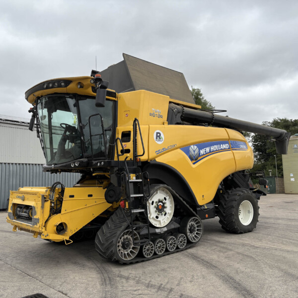 Yellow New Holland combine harvester on a paved lot, with a gray overcast sky and industrial buildings in the background.