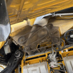 Close-up of a yellow construction machine interior showing gears, chains, belts and hydraulic components in a dusty, industrial setup.