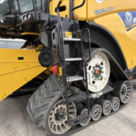 Close-up of a yellow New Holland construction machine showing its black undercarriage, steps, and multiple metal track rollers.