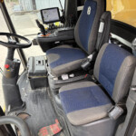 Cab interior of heavy equipment showing a blue upholstered driver seat, control joystick, and a mounted display screen beside the steering wheel.