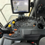 Inside a tractor or heavy machinery cabin, showing a large multifunction display screen above a curved control panel with multiple joysticks, yellow knobs, and numerous labeled buttons.