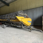 Yellow agricultural header/harvester inside a metal shed, with large grain bags labeled 'WYNNST' nearby