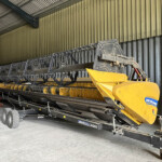 Yellow agricultural combine header inside a storage shed with metal walls and wheels visible beneath the frame.