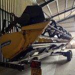 Yellow New Holland combine harvester stored inside a metal shed with a long grain header and racks along the wall.
