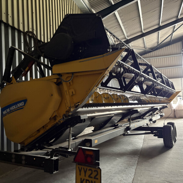 Yellow New Holland combine harvester stored inside a metal shed with a long grain header and racks along the wall.