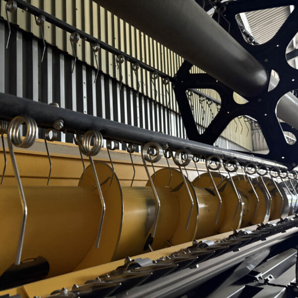 Close-up of an industrial machine showing a yellow support beam, a row of spiral springs along a black rail, and a dark blue structural frame in the background.