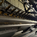 Close-up of a yellow conveyor frame with black metal brackets, springs, and a row of angled metal fingers along a dark industrial machine trail.