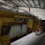 Yellow industrial machine inside a metal-walled workshop, with a long conveyor and row of hanging hooks along its top edge.
