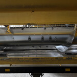 Underbelly of yellow industrial equipment with metal brackets, riveted panels, and fasteners visible in a workshop.