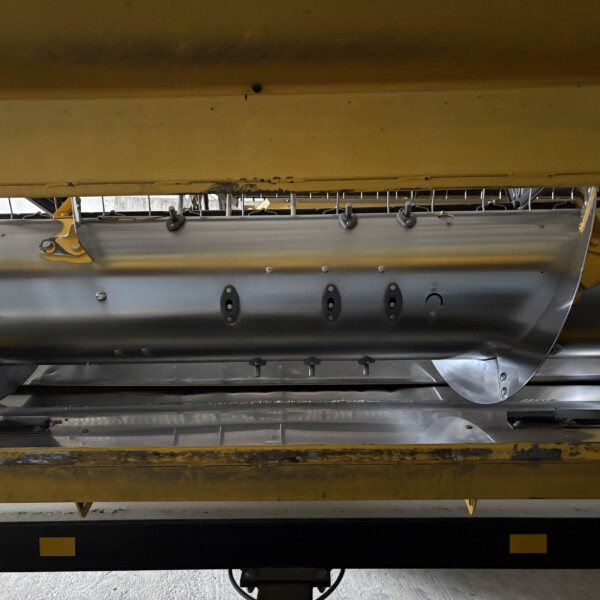 Underbelly of yellow industrial equipment with metal brackets, riveted panels, and fasteners visible in a workshop.