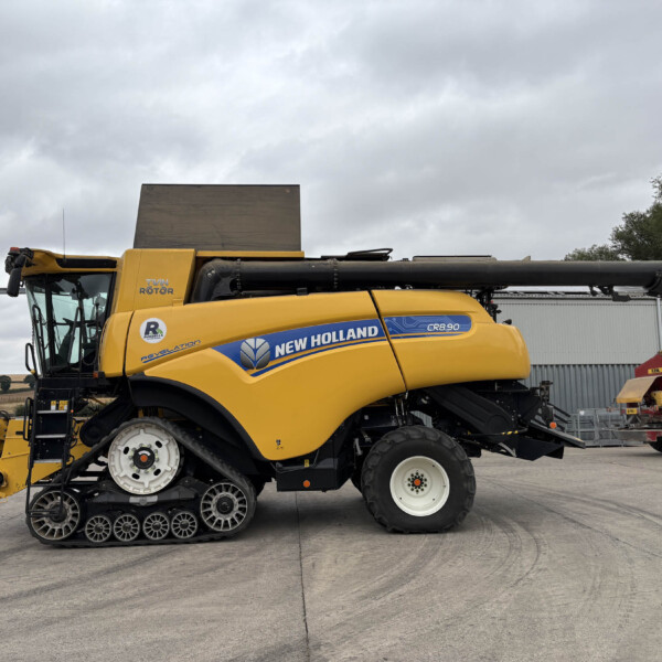 Side view of a yellow New Holland CR8.90 combine harvester with a large unloading auger, parked in a paved lot under a cloudy sky.