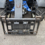 Rear view of a forklift attachment with rusty forks and a blue frame, on a paved surface outdoors.