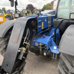 Close-up of a blue tractor chassis with hydraulic hoses and large front wheel arches, in a yard of construction equipment behind it.