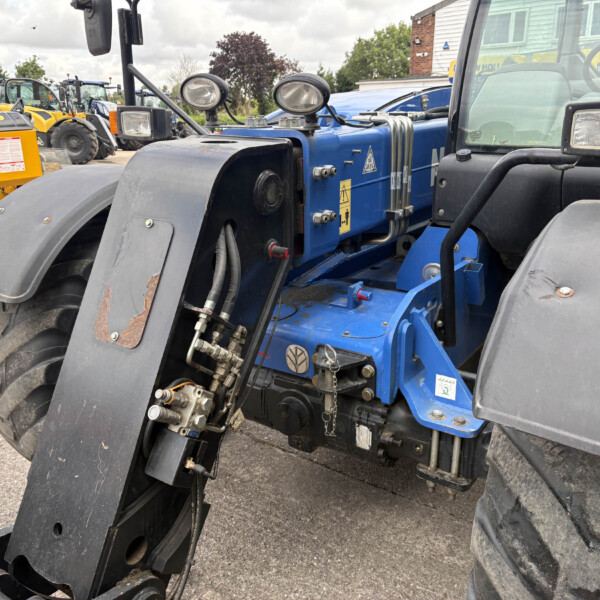 Close-up of a blue tractor chassis with hydraulic hoses and large front wheel arches, in a yard of construction equipment behind it.