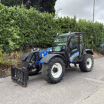 Blue New Holland tractor with a front-end loader parked along a street beside a tall green hedge under a cloudy sky, suburban setting.