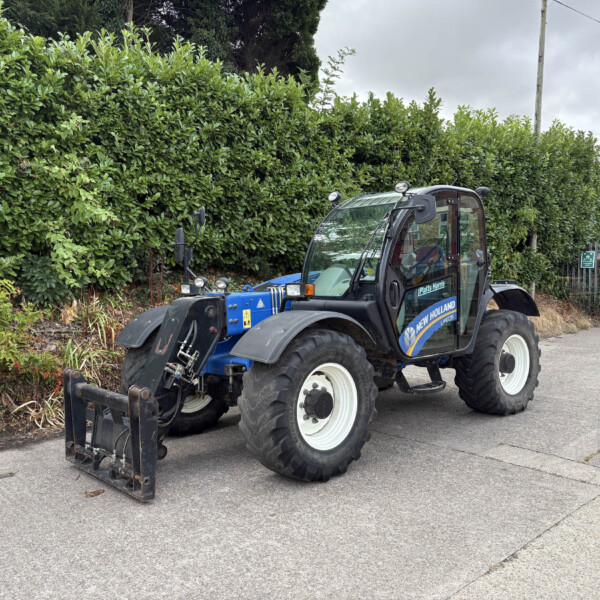 Blue New Holland tractor with a front-end loader parked along a street beside a tall green hedge under a cloudy sky, suburban setting.