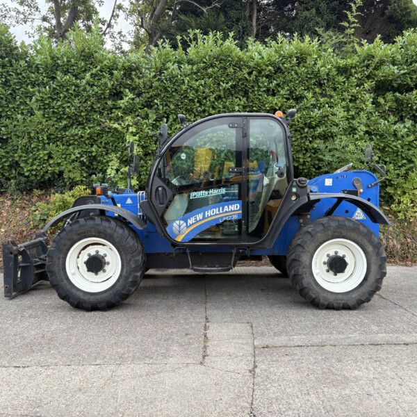 Blue New Holland tractor parked on a pavement with a tall green hedge behind it.