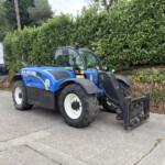 Blue New Holland tractor parked in a lot beside a tall green hedge, with a white building in the background.