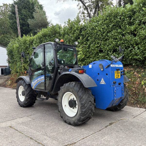 Blue New Holland tractor with front loader parked beside a tall green hedge near a white building with a yellow Sells sign in the background.