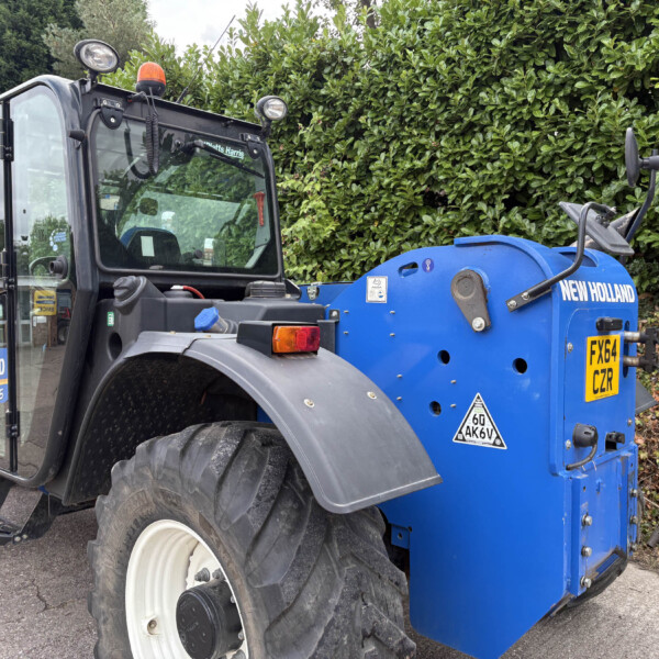 New Holland tractor with a blue attachment parked beside a hedge, showing the cabin and large front tire in a driveway setting