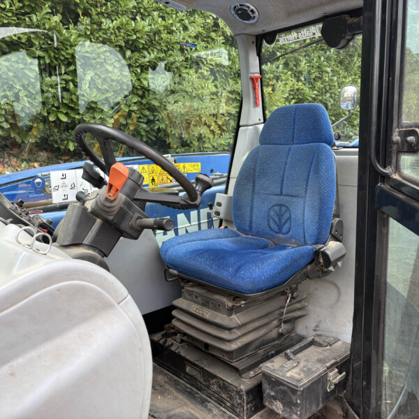 Interior of a blue tractor cab showing the steering wheel, seat, and control levers with yellow warning decals; outside greenery is visible through the window.