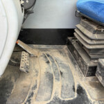 Dusty vehicle cab floor with pedals visible (accelerator and brake) and a blue seat on the right side.