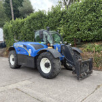 Blue New Holland tractor parked outdoors beside a tall green hedge with a building and a pickup truck nearby.