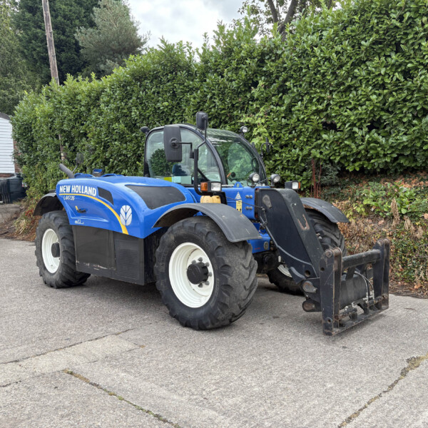 Blue New Holland tractor parked outdoors beside a tall green hedge with a building and a pickup truck nearby.