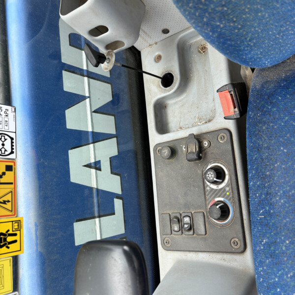 Inside a vehicle cab: blue exterior, control panel with levers, buttons, and a circular dial near a blue upholstered seat edge.