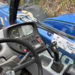 Inside a blue tractor cab showing the steering wheel, dashboard gauges, and control levers with hydraulic hoses nearby.