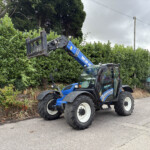 Blue New Holland construction loader with extended arm over a green hedge in an outdoor street setting. The vehicle is lifting the attachment near a hedge and sidewalk.