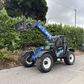Blue New Holland construction loader with extended arm over a green hedge in an outdoor street setting. The vehicle is lifting the attachment near a hedge and sidewalk.
