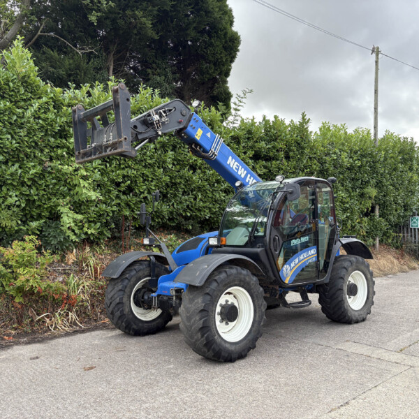 Blue New Holland construction loader with extended arm over a green hedge in an outdoor street setting. The vehicle is lifting the attachment near a hedge and sidewalk.