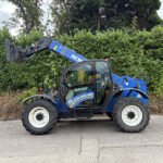 Blue New Holland front loader parked beside a tall green hedge, with the cab visible and raised bucket attachment awaiting use.