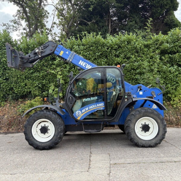Blue New Holland front loader parked beside a tall green hedge, with the cab visible and raised bucket attachment awaiting use.