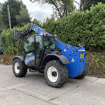 Blue New Holland telehandler with raised arm parked near a dense hedge and a white building in the background, construction site feel.
