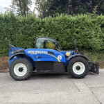 Blue New Holland compact tractor parked on a paved surface with a dense green hedge behind it, outdoors.