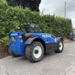 Blue New Holland tractor parked beside a tall green hedge on a street with overcast sky and red brick houses in the background
