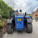 Rear view of a blue New Holland tractor parked beside a hedge in a residential area.