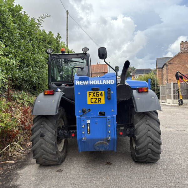 Rear view of a blue New Holland tractor parked beside a hedge in a residential area.