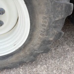 Close-up of a dirty white wheel rim with three rusted lug nuts visible and the thick black tire beside it on gravel ground.