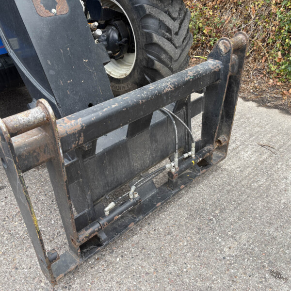 Rusty fork attachment for heavy machinery with hydraulic hoses, set on asphalt near ivy-covered ground.