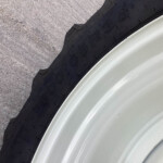 Close-up of a black tire with visible tread next to a light-colored metal rim on rough pavement surface.