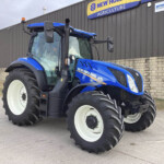Blue New Holland tractor parked on a concrete lot near a gray building with a yellow and blue New Holland Agriculture sign above.