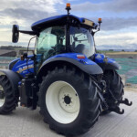 Blue New Holland tractor at a construction site, featuring large white wheels and a glass cab, viewed from the front-left.