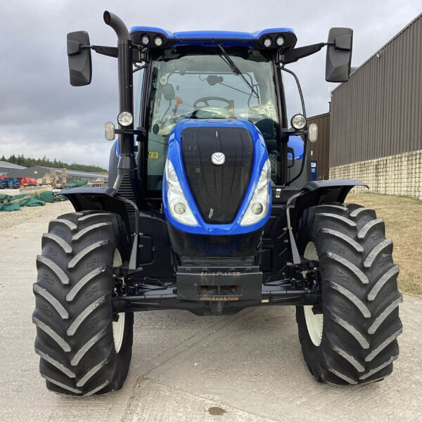 Front-on view of a blue agricultural tractor with oversized treaded tires, facing the camera in an industrial yard.