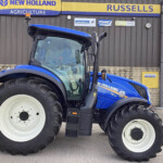 Blue New Holland tractor with oversized front tires parked in front of a building displaying New Holland Agriculture signage.