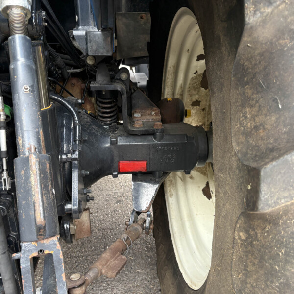 Close-up of a tractor's front hydraulic system and hitch, with rusted metal brackets and a large treaded tire nearby.