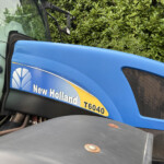 Close-up of a blue New Holland tractor with a visible T6040 model label and cab, hedge in the background.