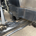 View under the front of a dirty black truck chassis with hoses, brackets, and rusted metal frame exposing wear and grime.